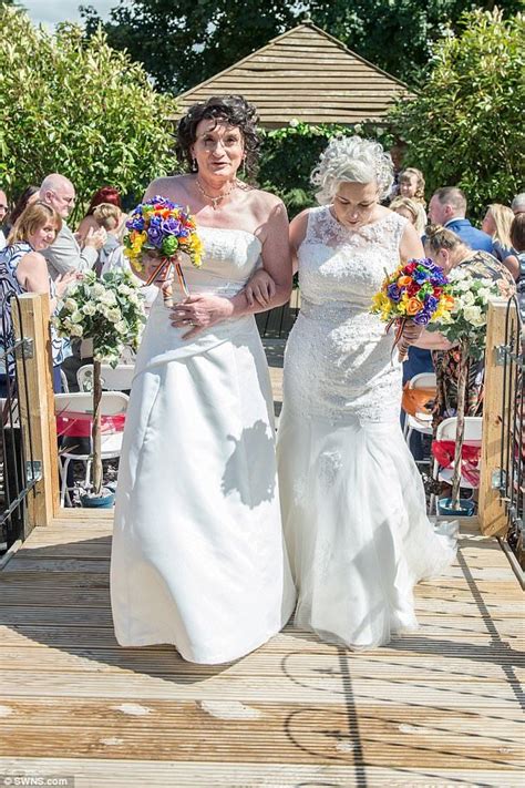 Two Women In White Dresses Walking Down A Wooden Walkway With Flowers