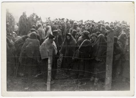 Survivors Wearing Blankets For Warmth Crowd Together Behind A Barbed