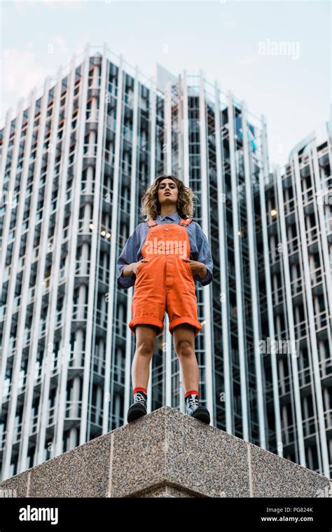 beautiful woman wearing dungarees standing on ledge in front of modern high rise building stock
