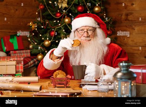 Hungry Santa In Costume Sitting By Table And Eating Cookies With Milk