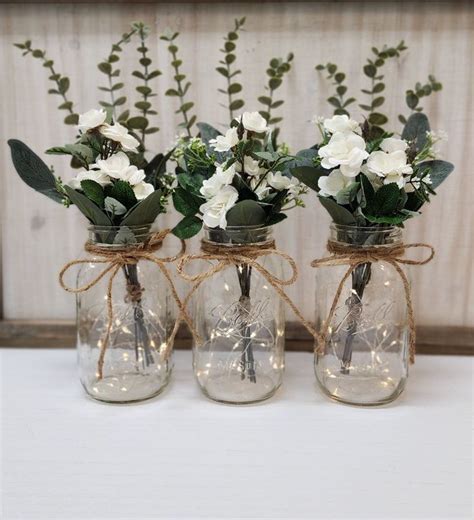 Three Mason Jar Vases With White Flowers And Greenery Tied Around The Top Sitting On A Table