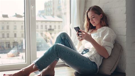 Beautiful Young Woman Sitting On A Windowsill At Home And Texting On