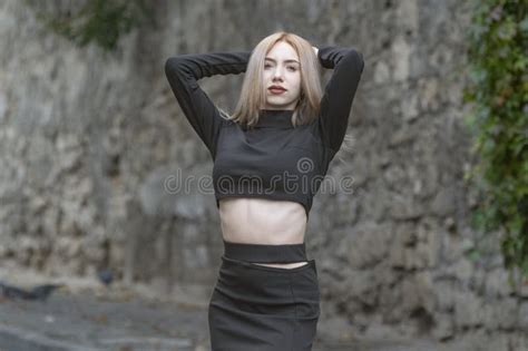 Slender Blonde Woman Wears Black Tight Fitting Clothes With Her Hands Behind Head And Looks Into