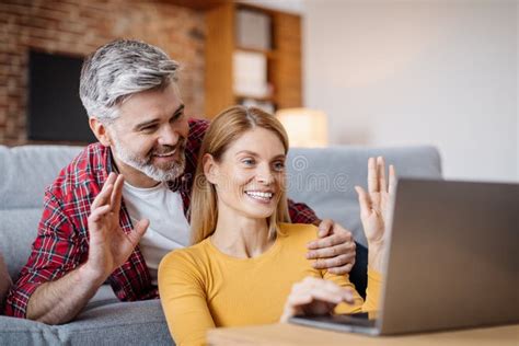 Glad Smiling Middle Aged Caucasian Husband Embracing Wife Waving Hand In Laptop Making Online