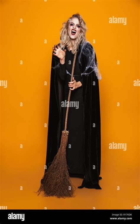 Angry Woman Witch With Scary Make Up Looking Camera And Holding Broom Isolated Over Orange Stock
