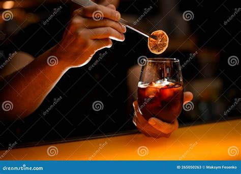 Hand Of Bartender Neatly Holds Glass With Cocktail And Garnishes It With Slice Of Dry Orange