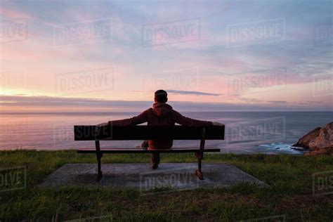 Full Length Of Man Looking At Sea While Sitting On Bench Against Cloudy