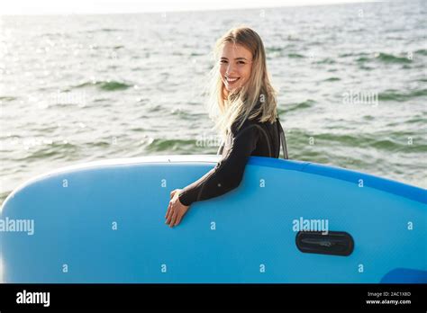 Image Of Smiling Blond Woman In Wetsuit Holding Surfboard While Walking On Summer Beach Stock