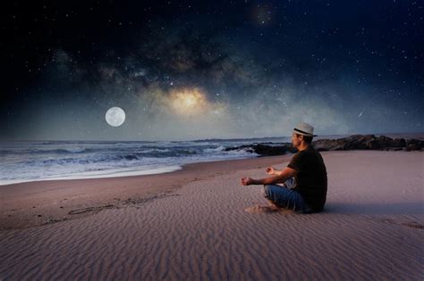 Человек сидит на пляже в позе лотоса медитируя на фоне млечного пути и луны Премиум Фото