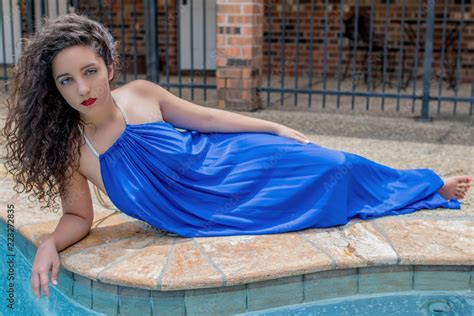 A Beautiful Tan Brunette Woman Lays In A Radiant Colored Blue Dress Beside A Teal Pool Outside