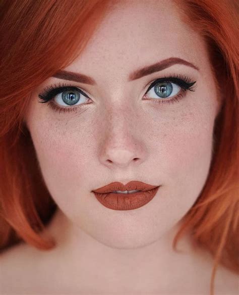 A Woman With Red Hair And Blue Eyes Wearing Orange Lipstick Looks At The Camera While Posing For