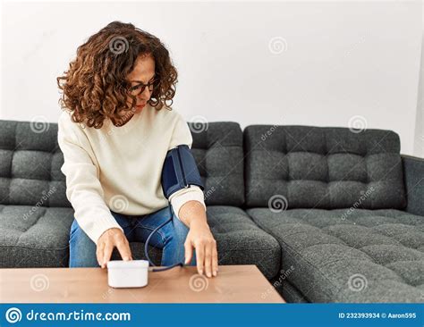 Mature Hispanic Woman Using Blood Pressure Monitor Sitting On The Sofa