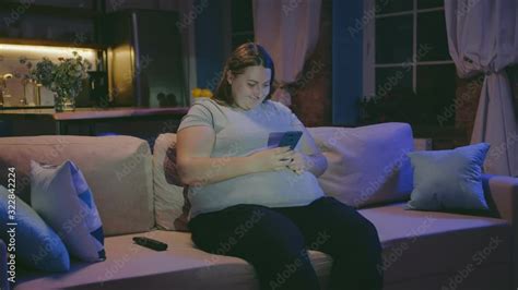 Overweight Woman Surfing Internet On Mobile Phone And Watching Tv Late