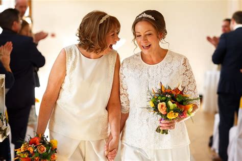 Two Brides Walking Down The Aisle Holding Hands And Smiling At Each