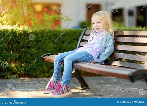 Cute Little Girl Sitting On Wooden Bench In A City Park On Summer Day