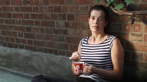 a woman sitting against a brick wall with a cup in her hand and looking at the camera