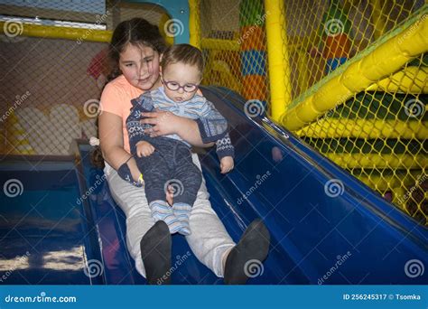 In The Playroom On The Slide The Sister Holds Her Little Brother In Her Arms And Slides Down