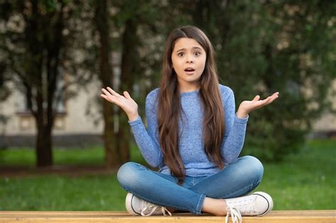 Шокированная девочка подросток пожимает плечами сидя скрестив ноги на скамейке на открытом