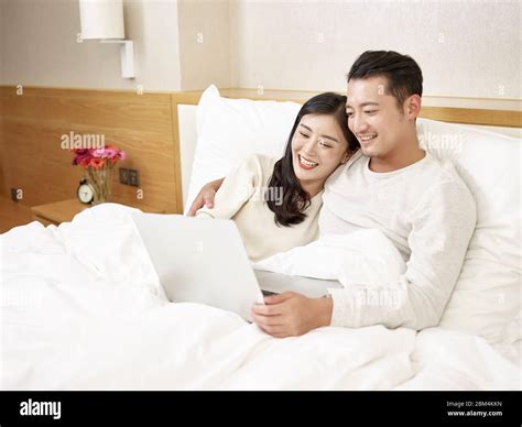 Young Asian Couple Sitting In Bed Enjoying A Movie Using Laptop