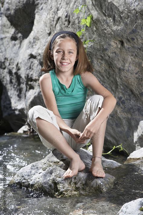 Full Length Portrait Of A Smiling Babe Girl Sitting On Rock By Stream