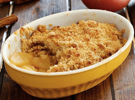 An apple crisp in a yellow dish on a wooden table next to apples and