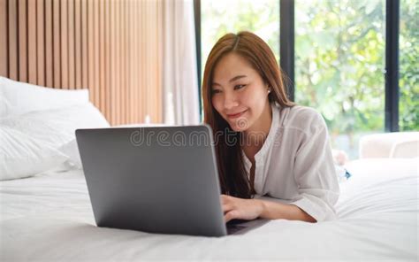 A Beautiful Woman Using And Working On Laptop Computer While Lying Down On A White Cozy Bed At
