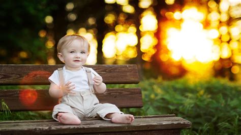 Cute Baby Boy Is Sitting On Wooden Bench Wearing White Dress In Blur