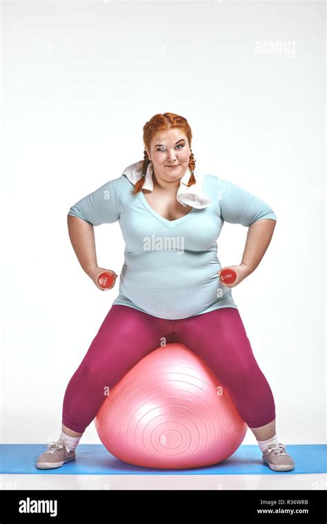 Red Haired Chubby Woman Is Sitting On The Ball And Keep The Dumbbells