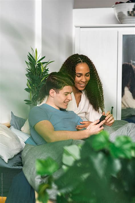 Interracial Couple Using Smartphone In Bed Del Colaborador De Stocksy