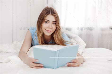 Beautiful Girl In Blue Pajamas Reading A Book Lying On The Bed In The