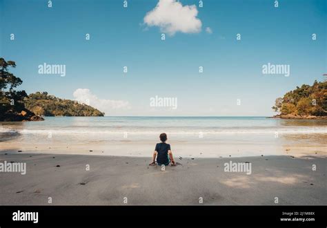 Boy Sitting Alone On Tropical Beach Looking At The Ocean On Sunny Day