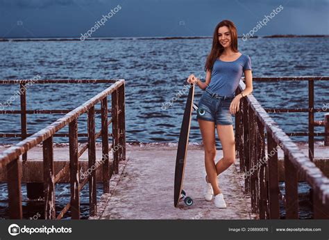 Sensual Girl Posing With Skateboard Standing On The Old Pier Is Enjoying Amazing Dark Cloudy