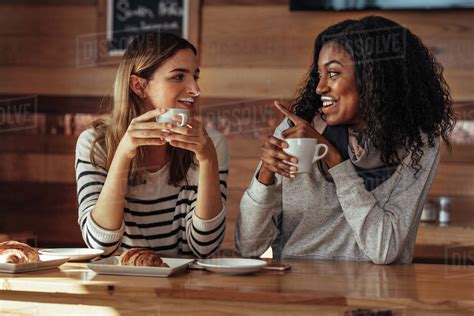 Two Women Drinking Coffee And Smiling At Each Other Pointing Towards Milk Mustache In Coffee