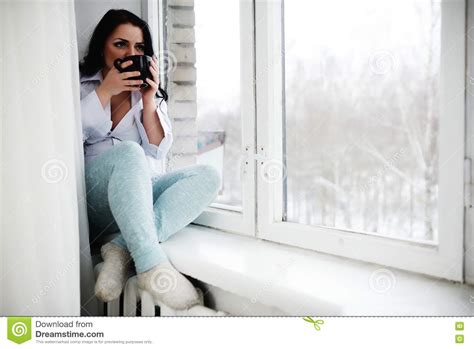 Молодая женщина сидит на силле окна с чашкой в руках и смотрит из окна Утро зимы Стоковое Фото