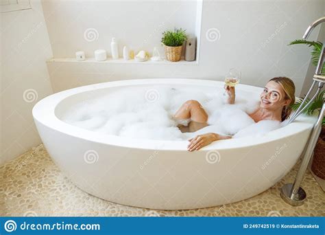 Blonde Beautiful Young Woman Lying In Bathtub With Foam And Holding Glass Of White Wine Stock