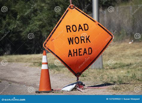 Battered Road Work Ahead Sign And Traffic Cone Sitting By Road With