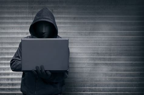 Premium Photo Hooded Hacker With Mask Holding Laptop While Typing