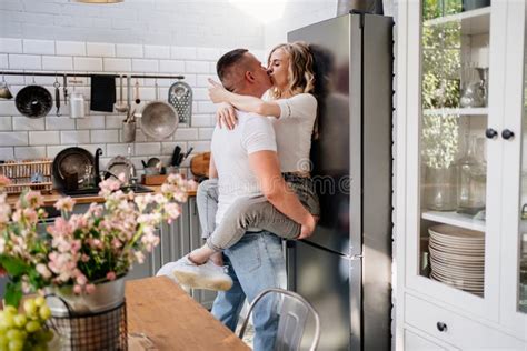 Pair Of Lovers Kissing And Hugging In Kitchen Next To The Refrigerator