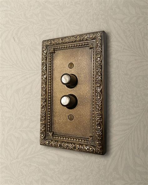 An Ornate Light Switch Plate With Two Dimmers On A Wallpapered Background