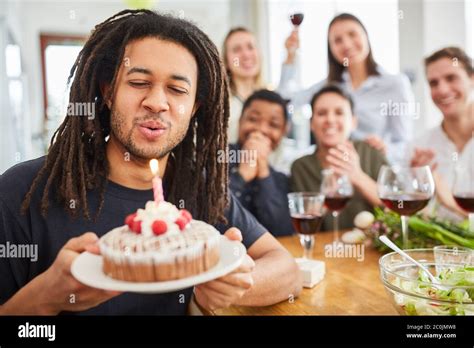 man with dreadlocks blowing out candle on cake at birthday party with friends at home stock