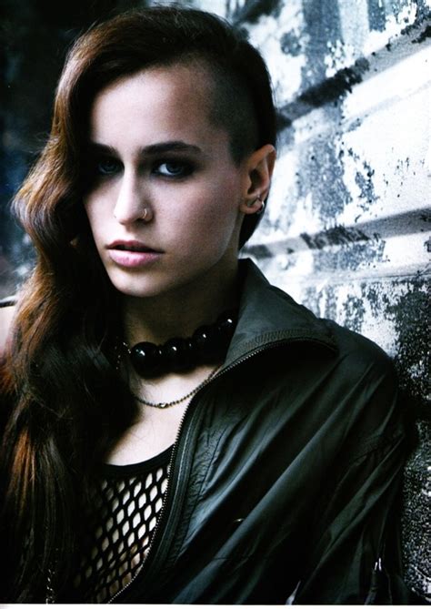a woman with long brown hair wearing a black leather jacket and choker leaning against a brick wall