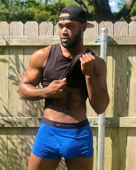 A Shirtless Man In Blue Trunks And A Cap Standing Next To A Wooden Fence