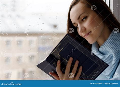 Young Brunette Woman In Blue Woolen Sweater Reading An Interesting Book On The Windowsill In