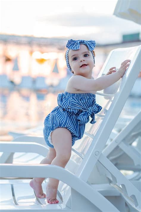 Baby Girl On Summer Vacation Standing On Lounger Next To Swimming Pool