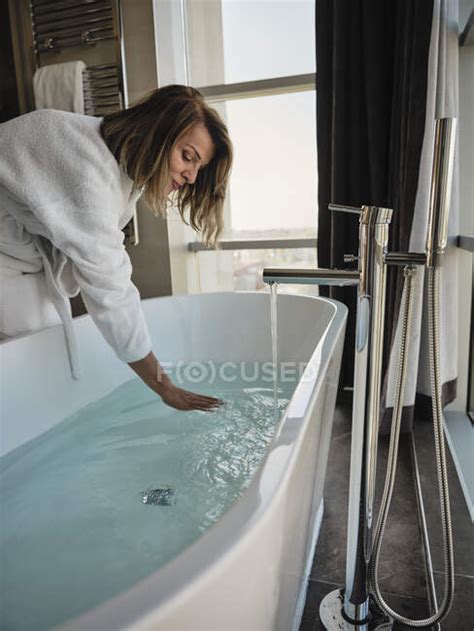 Senior Woman Bending Over While Examining Temperature In Bathtub At