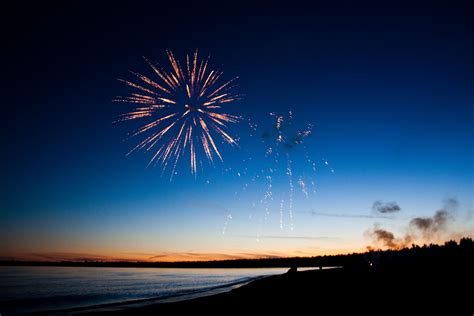 Fireworks Are Lit Up In The Night Sky Over The Water And Trees On The Shore