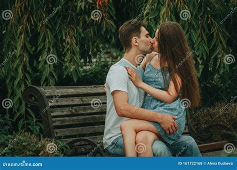 Beautiful Couple In Love Sits In A Park On A Bench In The Summer Hugs And Kisses The Girl Sits