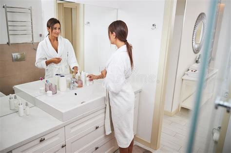 Brunette Woman In White Bathrobe Looking To The Reflection In The Mirror In Room Indoors Stock