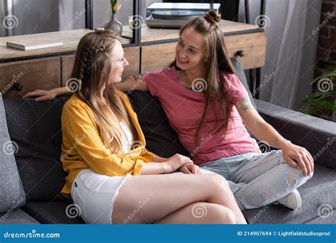 dos lesbianas sonrientes sentadas en el sofá y mirándose en el salón foto de archivo imagen
