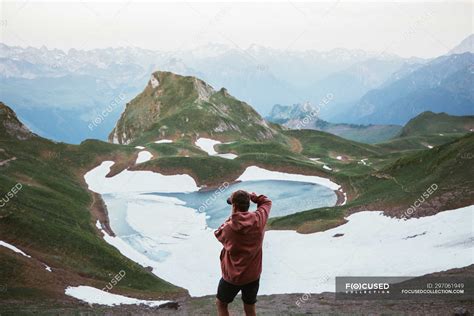 Турист фотографирует горное озеро окруженное снегом и хребтом в